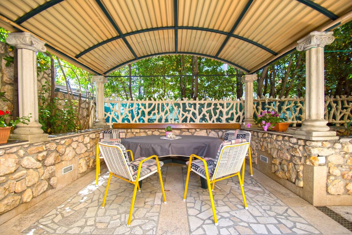 Ένας εξωτερικός χώρος με πέτρινα τοιχώματα, μια σκεπαστή περιοχή με καθίσματα και γλάστρες με φυτά.