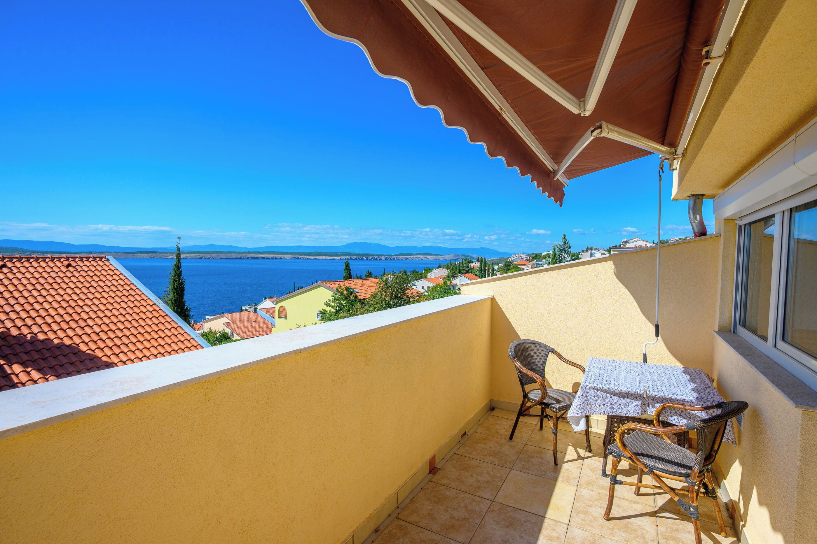 Ein Balkon mit Tisch und Stühlen, der Blick auf die Küste mit Häusern und einem klaren Himmel bietet.
