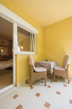 A room with a yellow wall, a small table with two wicker chairs, a window with a white frame, and a bed in the background. - Apartments Oaza (Dramalj)