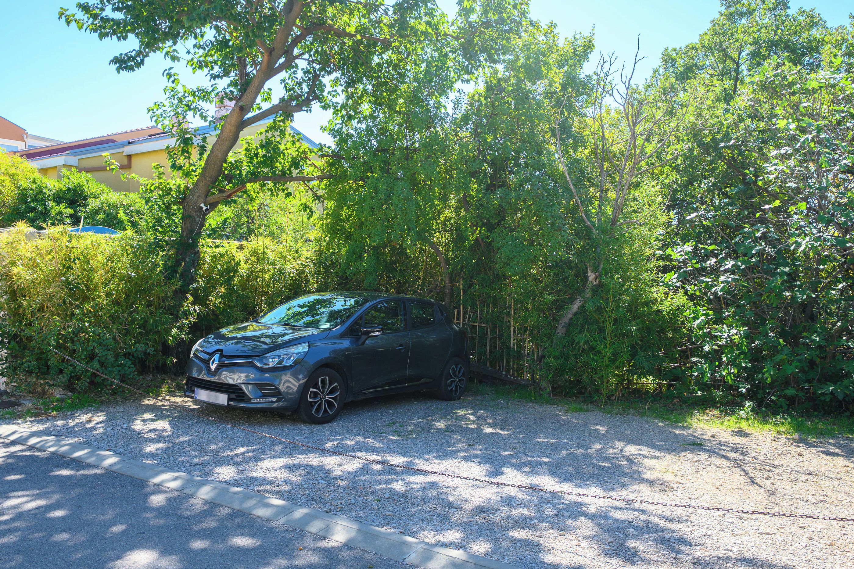 Carro estacionado em um caminho de cascalho com corrente bloqueando a entrada, rodeado por vegetação densa e árvores.