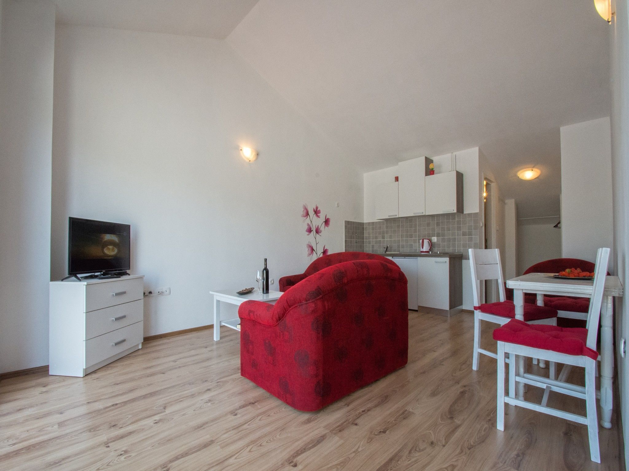 A compact living space with a kitchenette, a dining area with a red sofa and white chairs, and a television on a white cabinet.