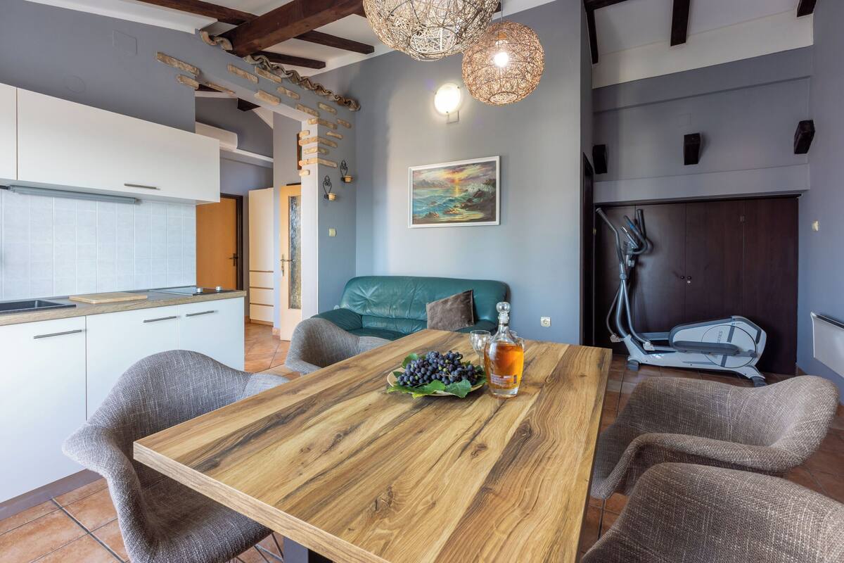 Een moderne keuken met witte kasten, een houten eettafel met een fles en druiven, en een grijze bank.