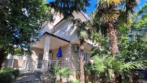 A stone building with a balcony, surrounded by tropical plants and a clear blue sky. - Apartments Danica (Dramalj)