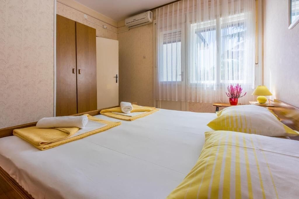 Hotellihuone, jossa on sänky, vaatekaappi, ikkuna verhoilla sekä pieni pöytä, jossa on lamppu ja maljakko.