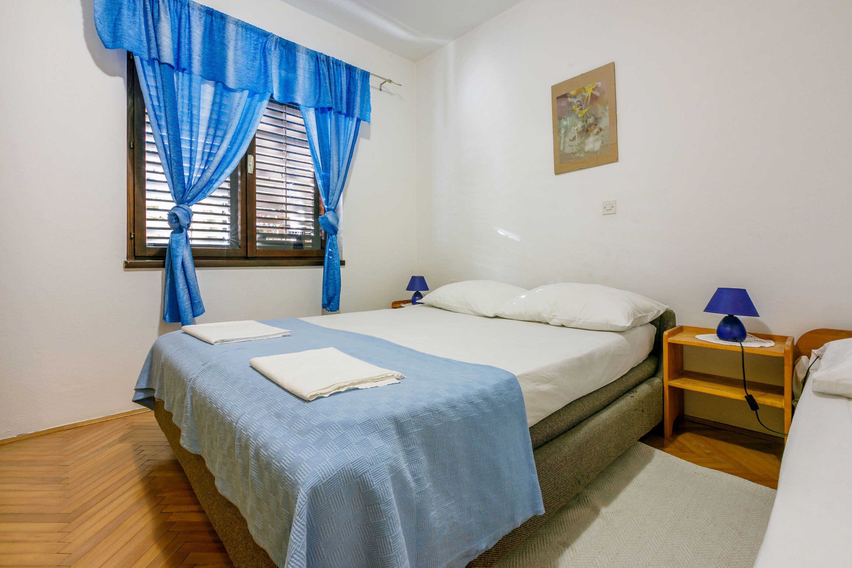 A bedroom with a bed, a window with blue curtains, a bedside table with a lamp, and a painting on the wall.