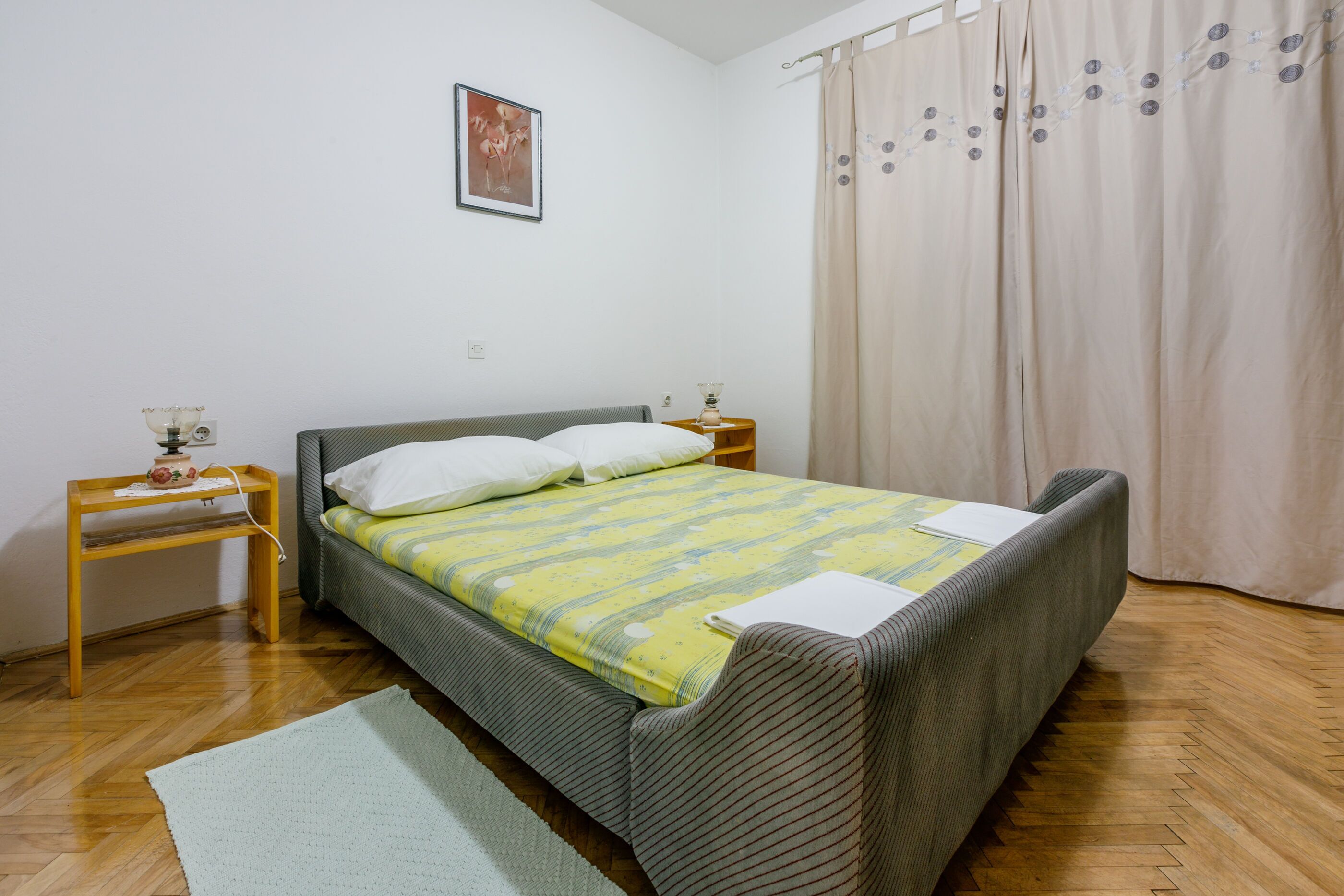 A bedroom with a bed, a nightstand, a lamp, a picture on the wall, and a window with curtains.