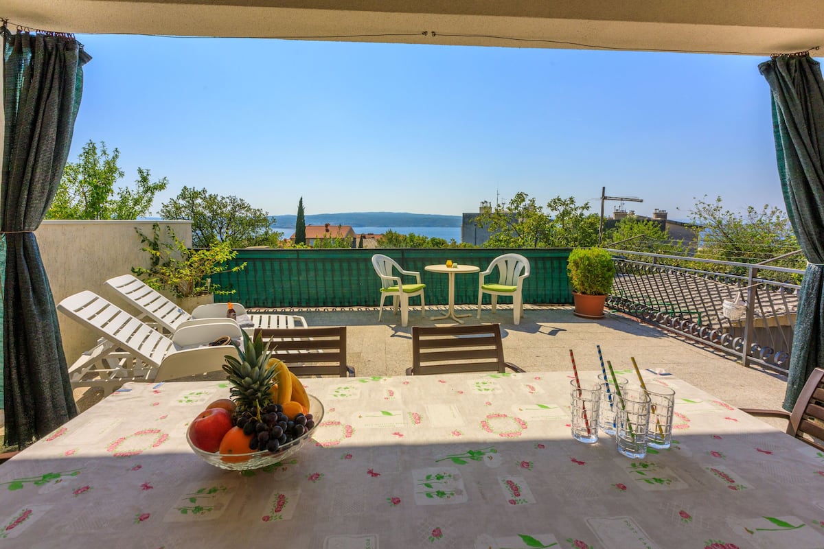 Une terrasse avec une table dressée pour le repas, des chaises, un saladier rempli de fruits et une vue sur la mer.