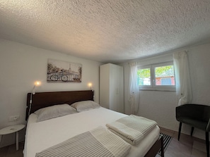 Une chambre à coucher avec un lit, une chaise, une fenêtre avec des rideaux, un luminaire fixé au mur et un tableau.