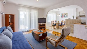 A modern living room with a sofa, a chair, a coffee table, and a TV. There is a dining area with a table and chairs. The room has a kitchen area with a counter and cabinets. - Apartments Alex (Crikvenica)