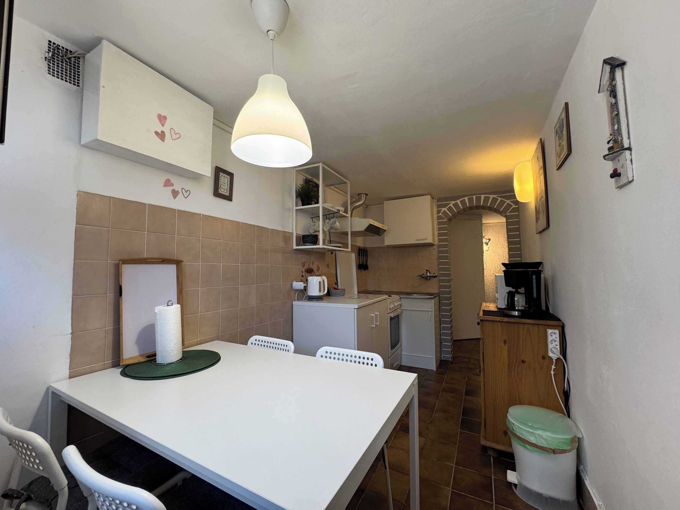 Cocina compacta con una mesa, sillas, lámpara colgante, cafetera y un cubo de basura.