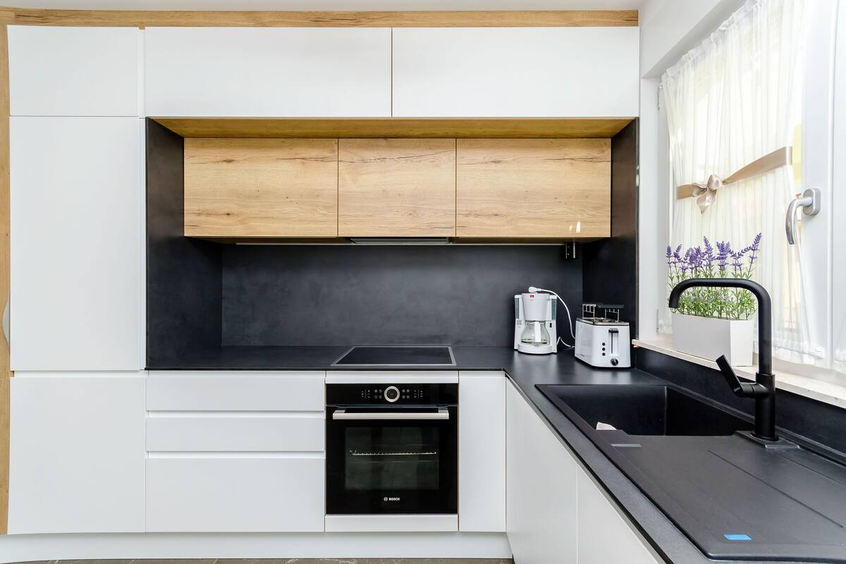 A modern kitchen with white cabinets, a black countertop, a Bosch oven, a coffee maker, and a window with a plant.