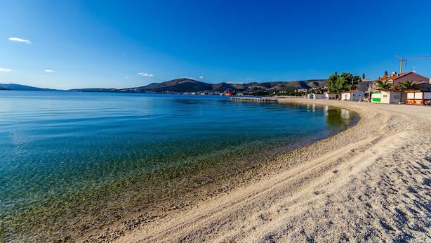 Μια παραλία με καθαρά νερά, αμμώδη ακτή και κτί ρια στο βάθος.