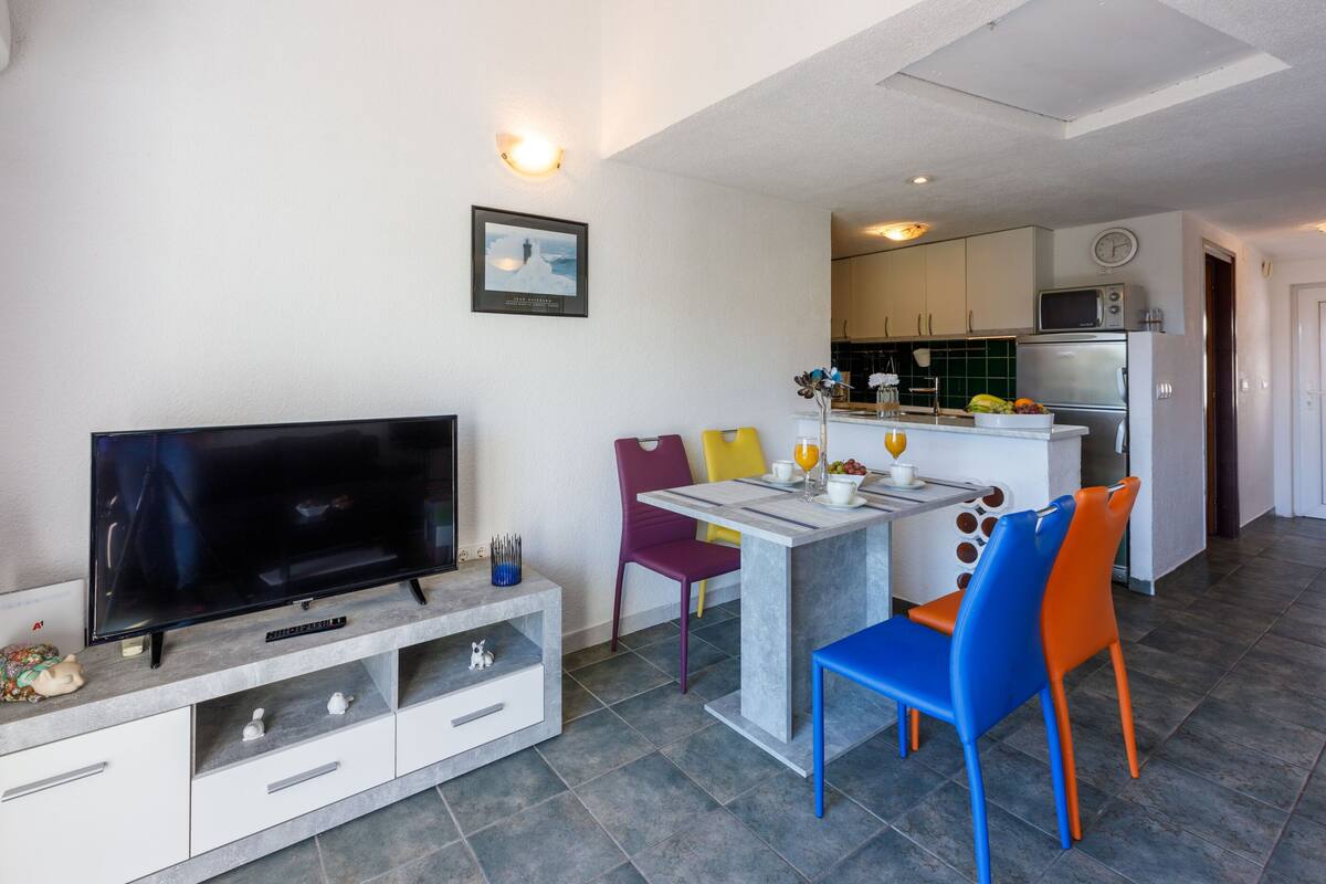 A modern living room with a flat-screen TV, a dining table with chairs, and a kitchen area with appliances.