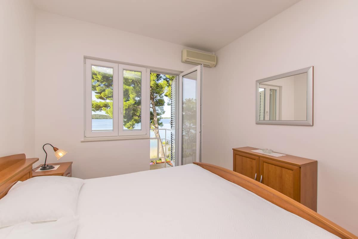 Un dormitorio con cama, mesita de noche, cómoda, ventana con vista a árboles y equipo de aire acondicionado.