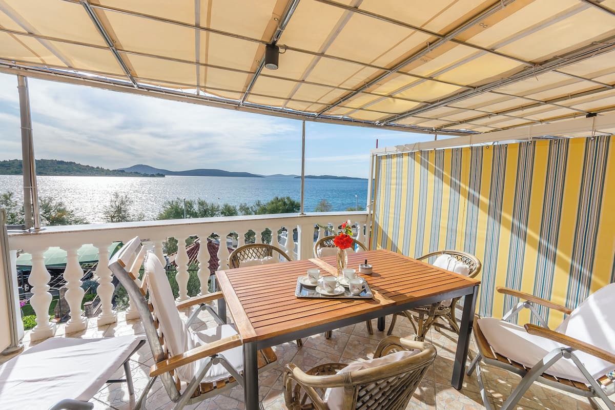 Eine Terrasse mit einem Holztisch, gedeckt für zwei Personen, Korbstühlen und Blick auf einen See und Berge.