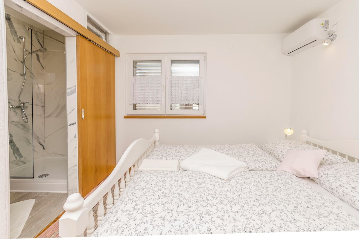 Ein Schlafzimmer mit Bett, Holzschrank, Duschbereich und einem Fenster mit Jalousien.