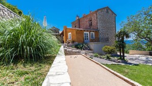 Une cour avec un terrain de pétanque, des bâtiments en pierre et une vue sur la mer.