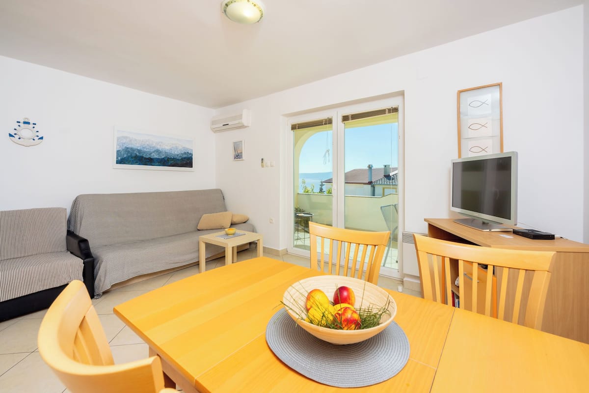 A modern living room with a sofa, a dining table with a fruit bowl, a TV, and a view of the sea through the glass door.