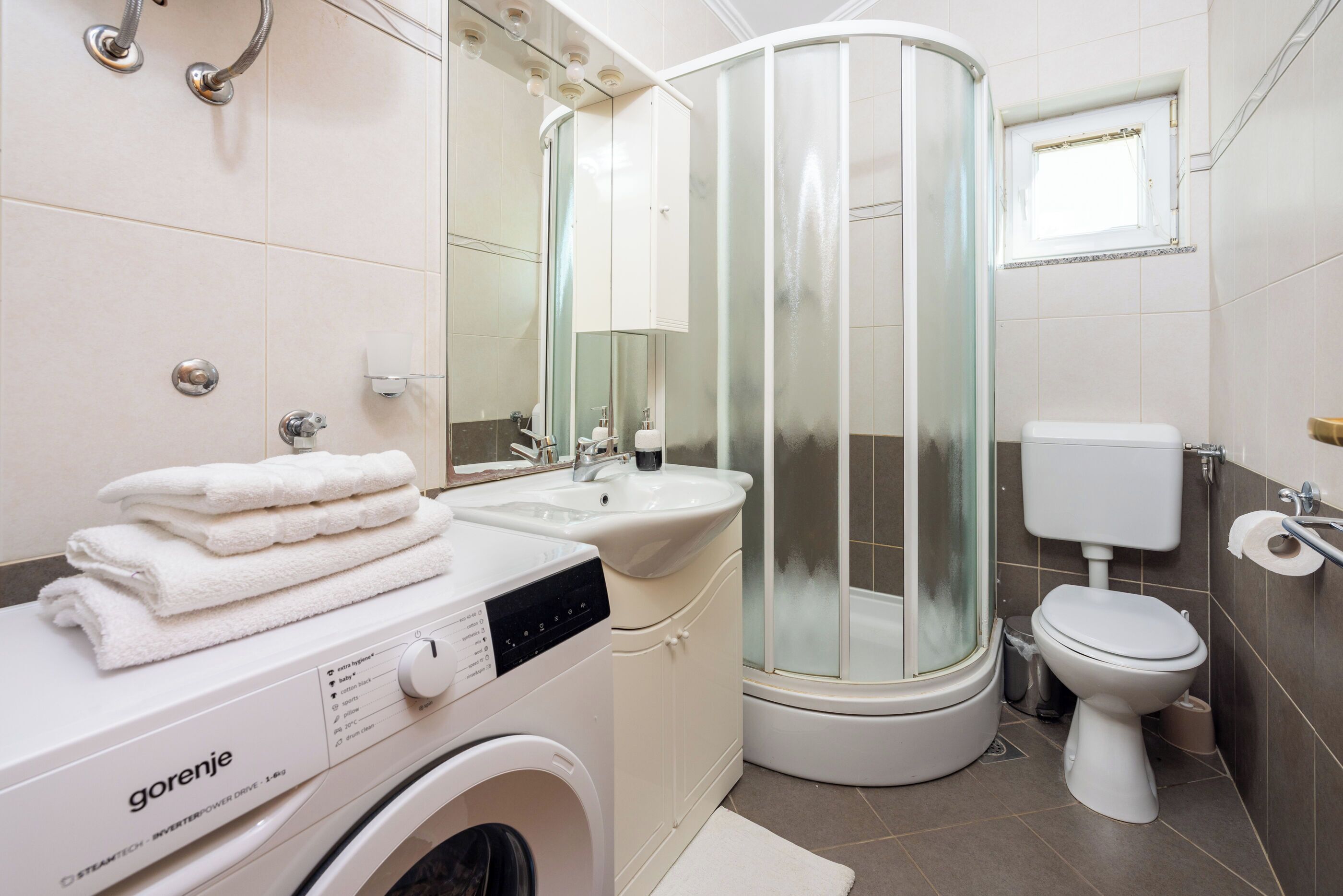 A modern bathroom with a washing machine, a glass-enclosed shower, a toilet, and a sink.