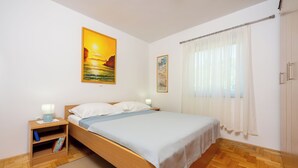 A neatly arranged bedroom with a wooden bed, a nightstand with a lamp, and a view of the outdoors through a window with sheer curtains. - Apartments Petak (Dramalj)