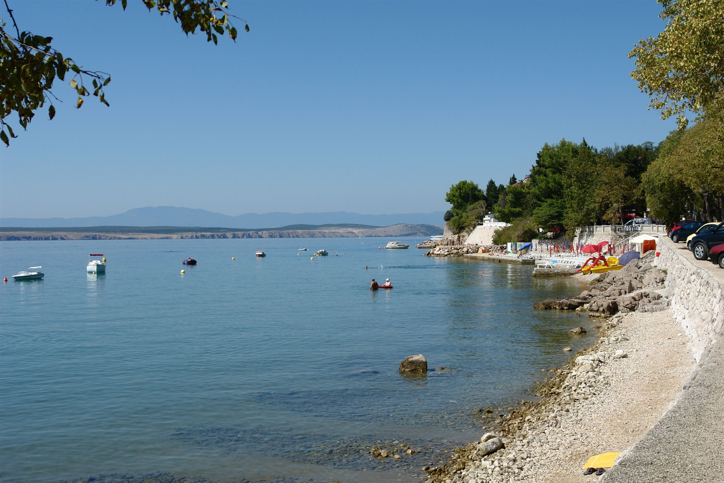 A coastal area with boats on calm waters, a rocky shoreline, and a clear sky.