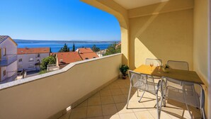 A balcony with a table and chairs, overlooking a coastal view with buildings and a clear sky. - Apartments Petak (Dramalj)