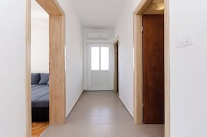 En smal hall med en trädörr på höger sida, en vit dörr längst ner i hallen och en säng med grått sänglinne på vänster sida.