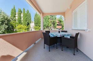 A balcony with a table and chairs, a view of trees and buildings, and a building with a red roof in the background. - Apartments Slavica (Lopar)