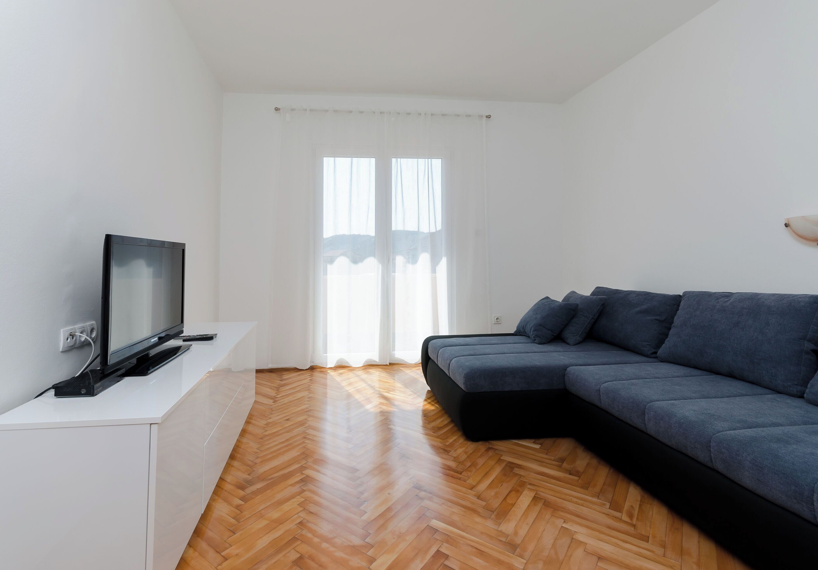 Un salon moderne avec un téléviseur à écran plat posé sur un meuble blanc, un canapé de couleur foncée et un parquet en bois à motifs chevrons.