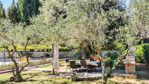 Ένας εξωτερικός χώρος με πλακόστρωτο δάπεδο, ένα τραπέζι και καρέκλες από πέτρα, περιτριγυρισμένος από δέντρα και έναν τοίχο από πέτρα.