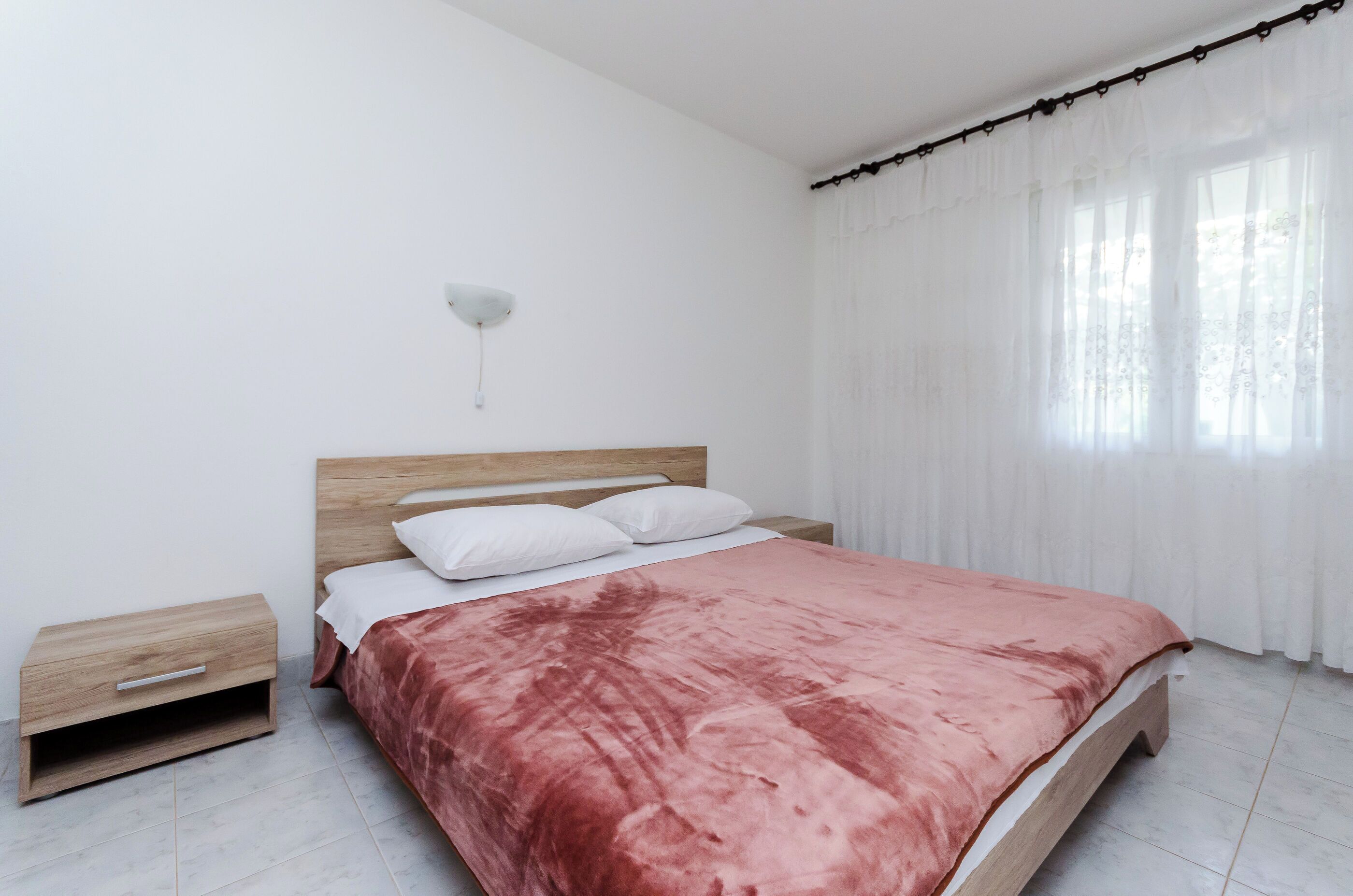A bedroom with a wooden bed frame, a pink bedspread, white pillows, a nightstand, and a wall-mounted light.