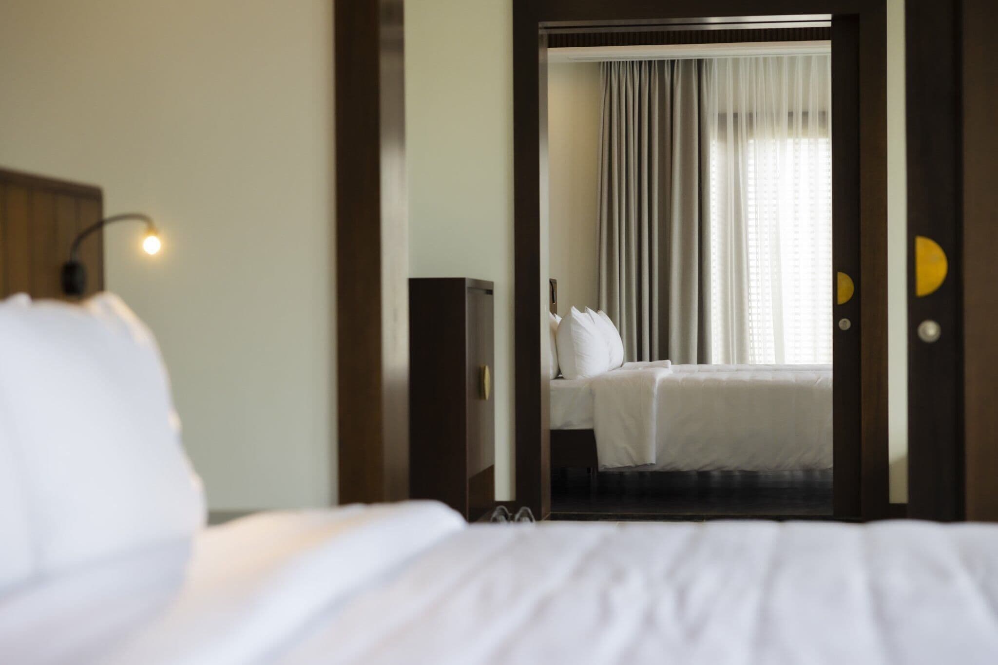 A hotel room with a bed, a nightstand, a lamp, and a view of another room through a door.