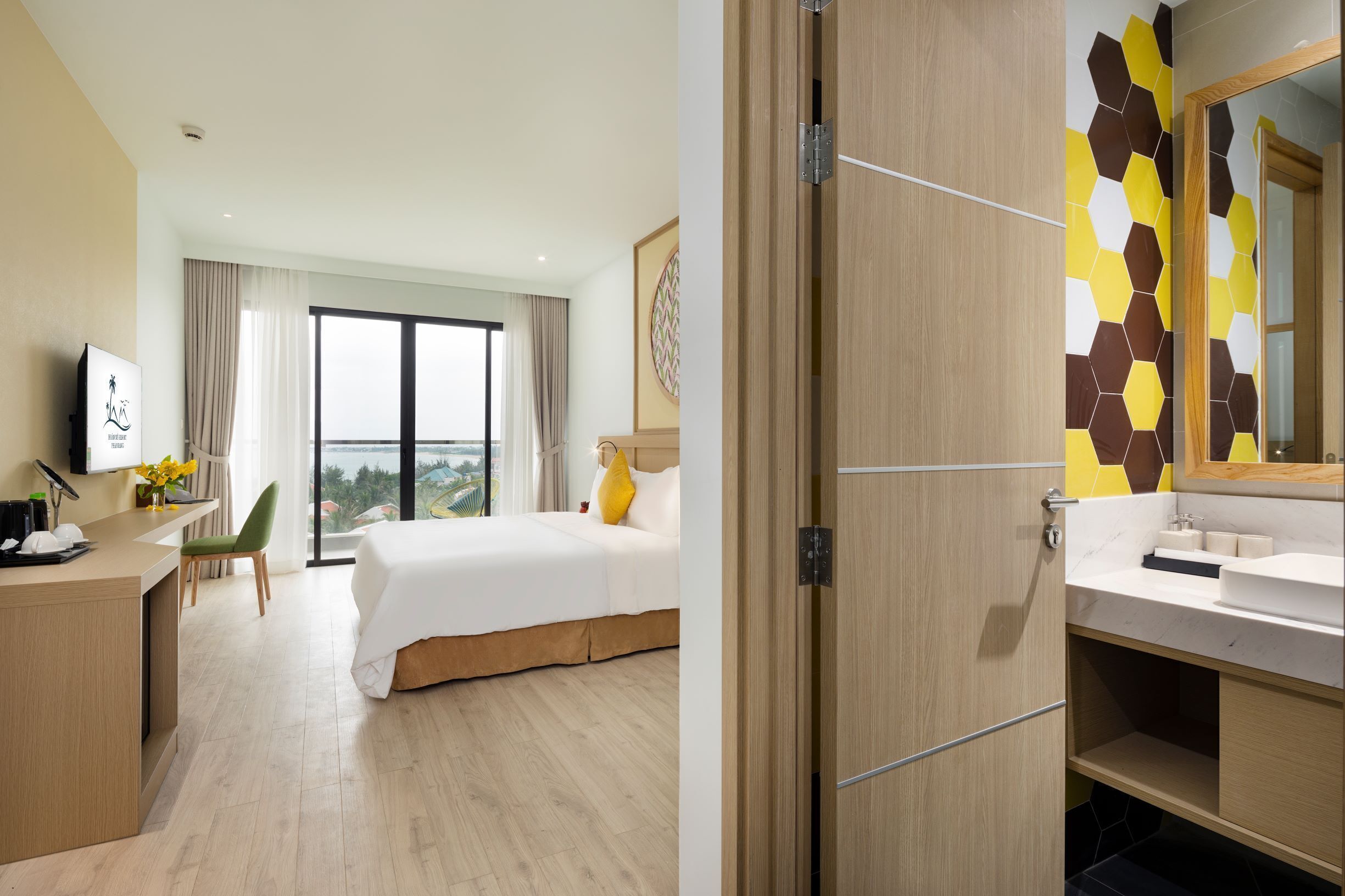 A hotel room with a bed, a desk, a chair, a large window, and a bathroom with a mirror and sink.