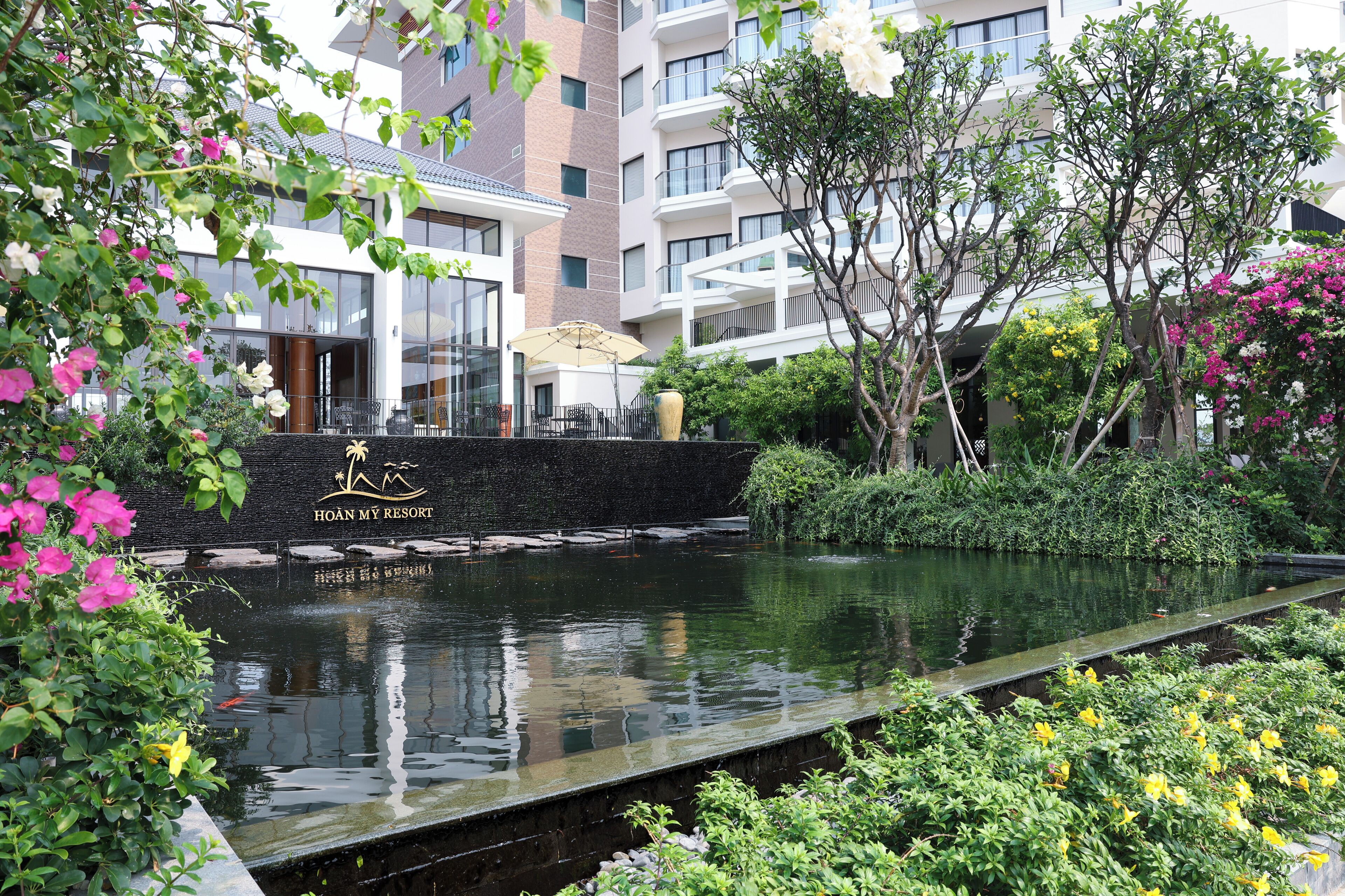 A serene hotel pool area with a stone wall, lush greenery, and a clear view of the hotel building in the background.