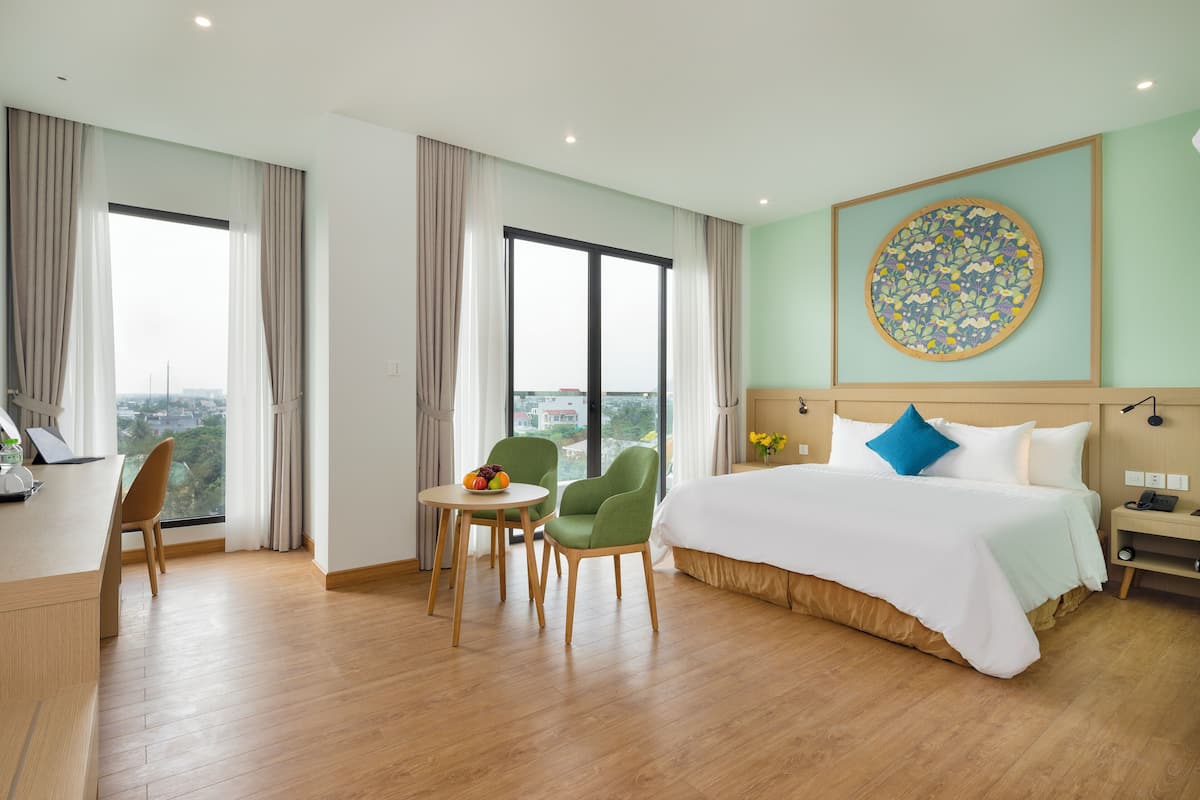 A hotel room with a large bed, a desk with a chair, a small table with fruit, and a view of the city through large windows.