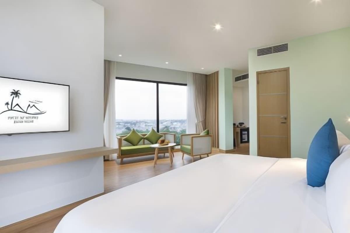 A hotel room with a large bed, a sofa, a small table, and a view of the city through large windows.