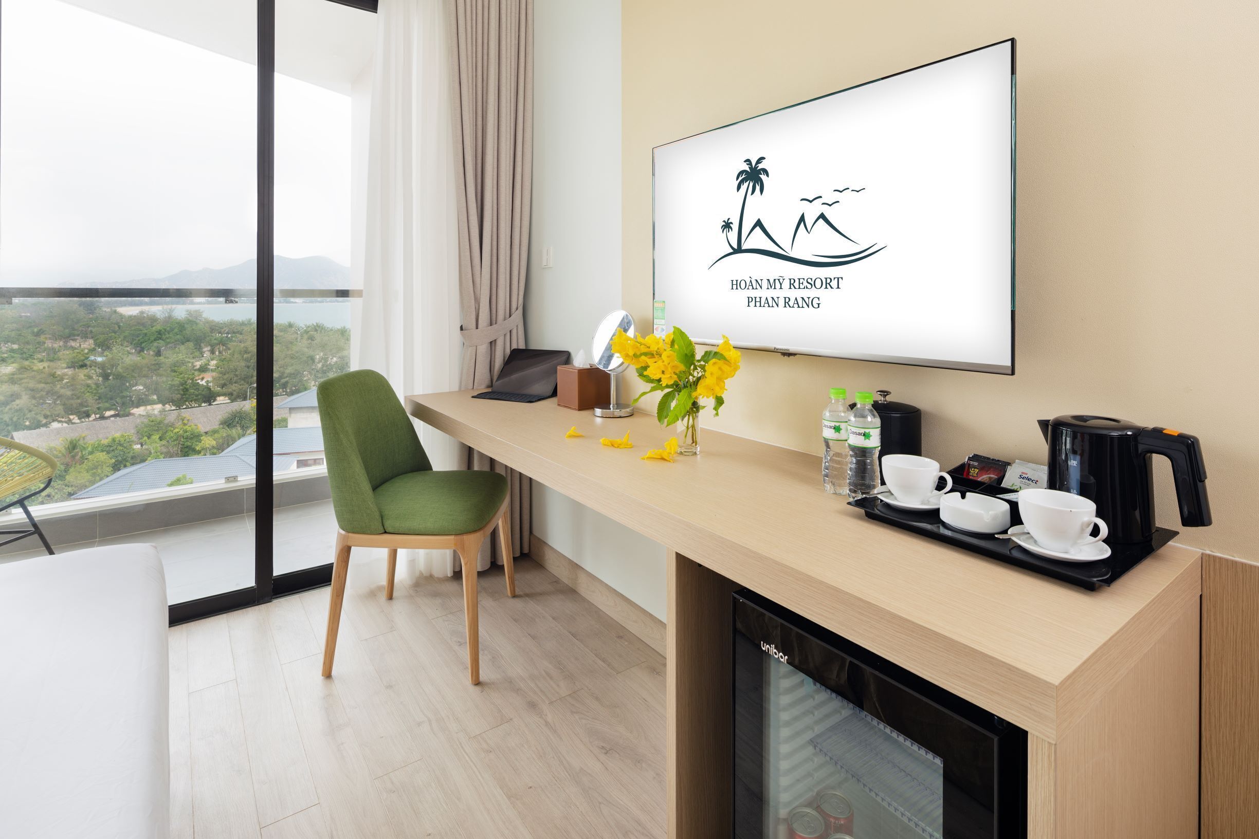 A modern hotel room with a balcony, a desk with a coffee maker, cups, and a vase with flowers.