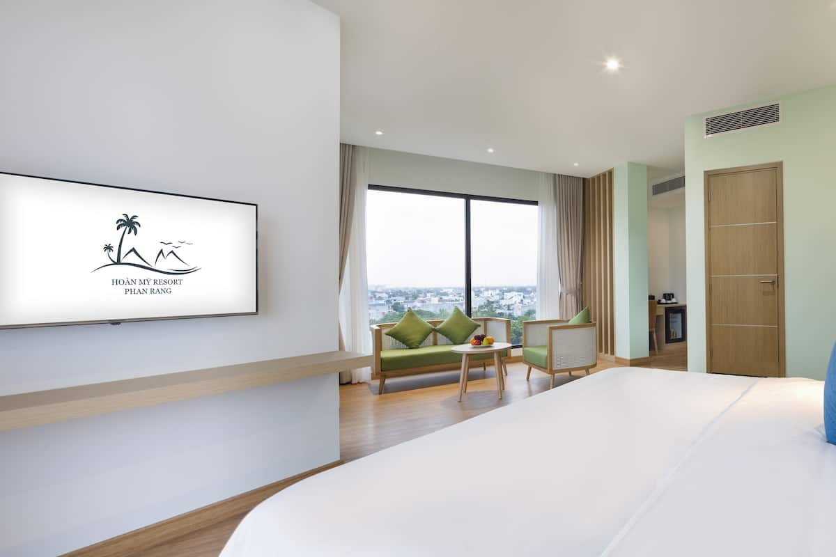 A modern hotel room with a large bed, a sofa, a small table with fruit, a desk, and a view of the city.