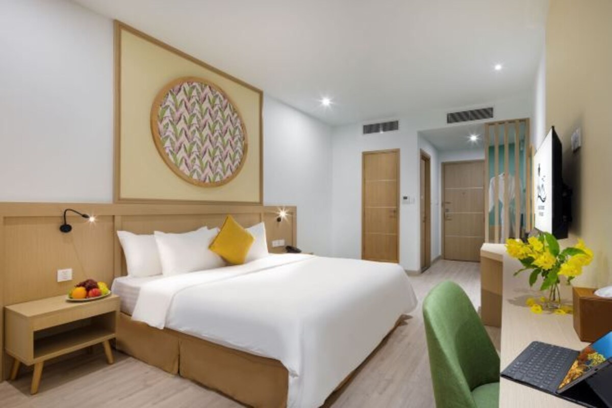 A hotel room with a bed, bedside table with fruit, a desk with a laptop, and a wall-mounted TV.