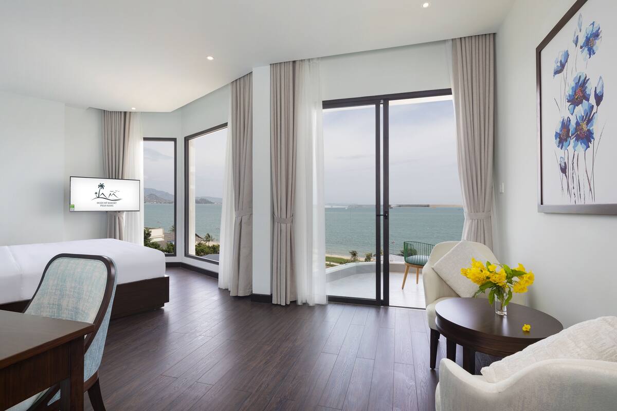 A modern hotel room with a bed, a desk, a chair, a TV, and a view of the sea.