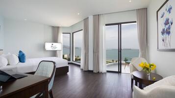 A modern hotel room with a bed, a desk, a chair, a TV, and a view of the sea.