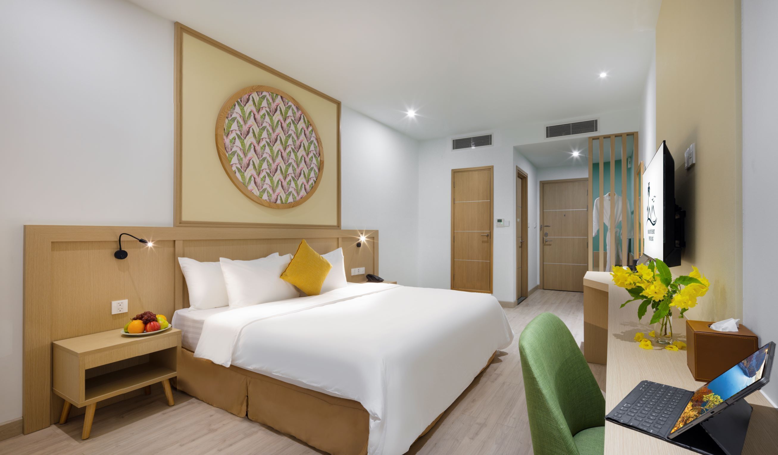 Une chambre d’hôtel avec un lit, une table de chevet avec des fruits, un bureau avec un ordinateur portable, une chaise verte, une télévision, et une salle de bain visible au fond.