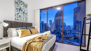 A bedroom with a large window offering a city view, a bed with a gold throw blanket, and a nightstand with a lamp. - Savannah Penthouse (Broadbeach)