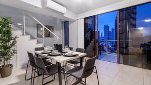 A modern dining area with a white table set for four, a staircase, and a city view through large glass doors. - Savannah Penthouse (Broadbeach)