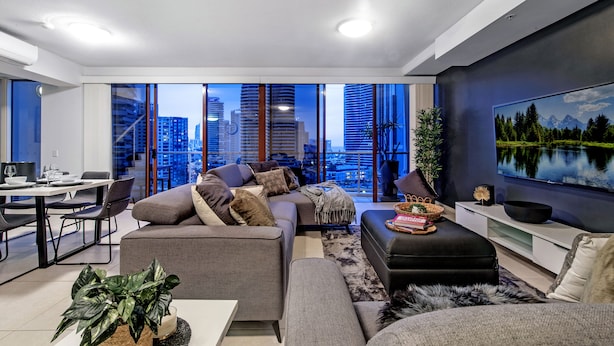 A modern living room with a city view, a dining area, and a flat-screen TV on the wall. - Savannah Penthouse (Broadbeach)