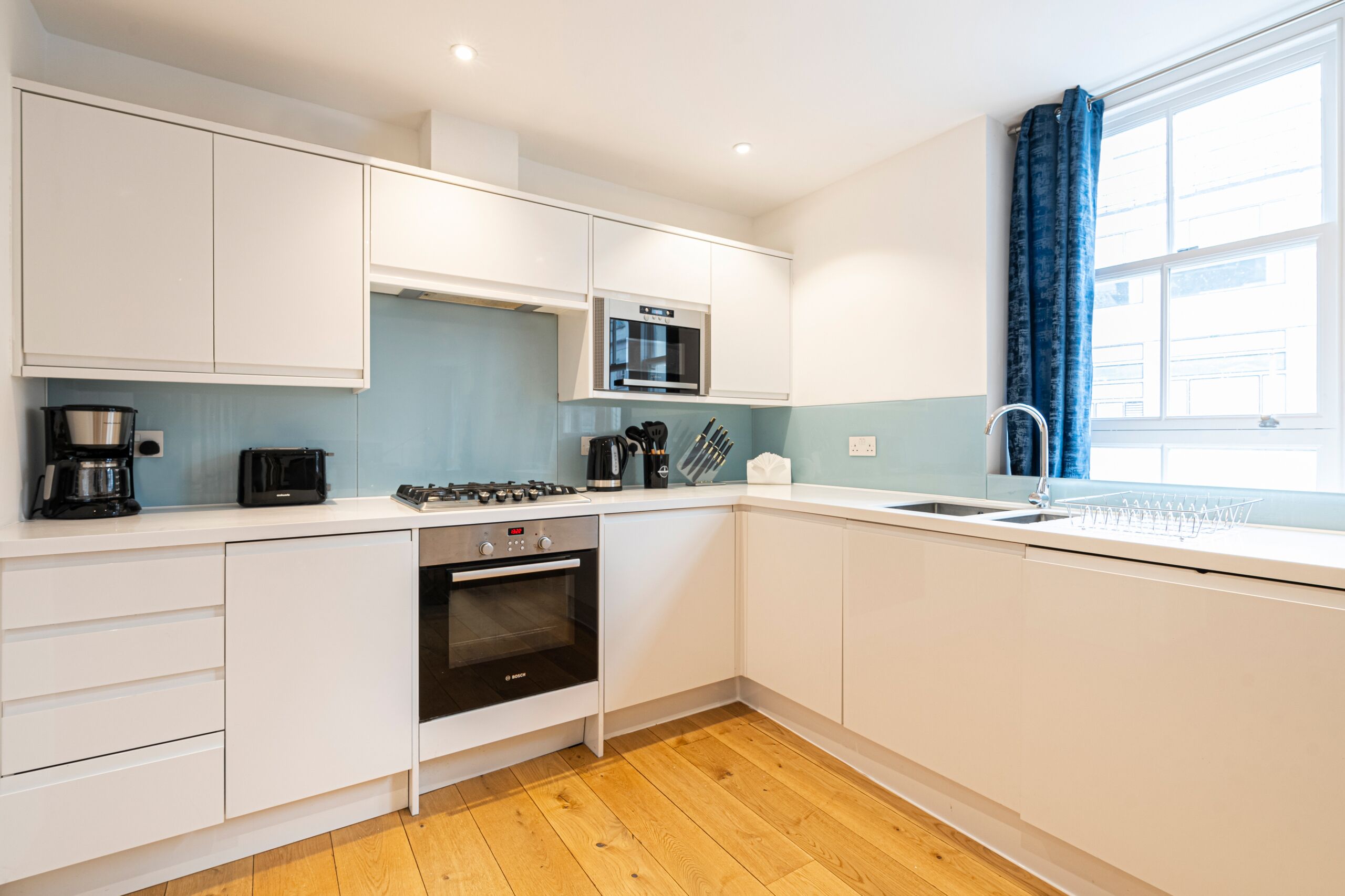 A modern kitchen with white cabinets, a built-in oven, a microwave, a coffee maker, and a window with blue curtains.