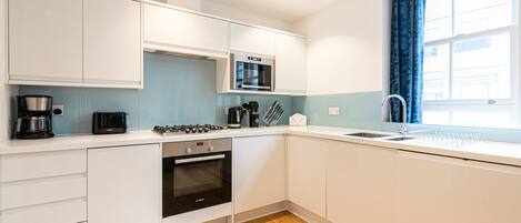 A modern kitchen with white cabinets, a built-in oven, a microwave, a coffee maker, and a window with blue curtains.
