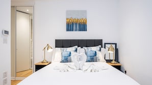 A hotel room with a large bed, two bedside tables with lamps, a wall-mounted artwork, and a door leading to another room. - Marylebone Apartments (London)