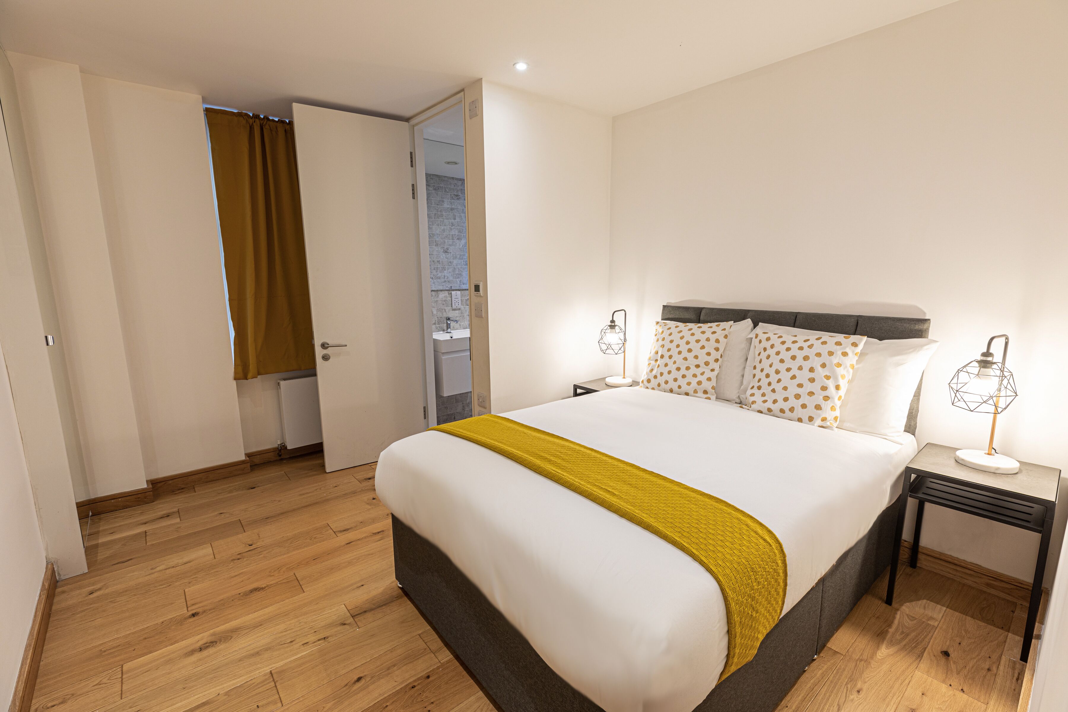 A neatly arranged bedroom with a large bed, a bedside table, a nightstand, and a bathroom visible through an open door.