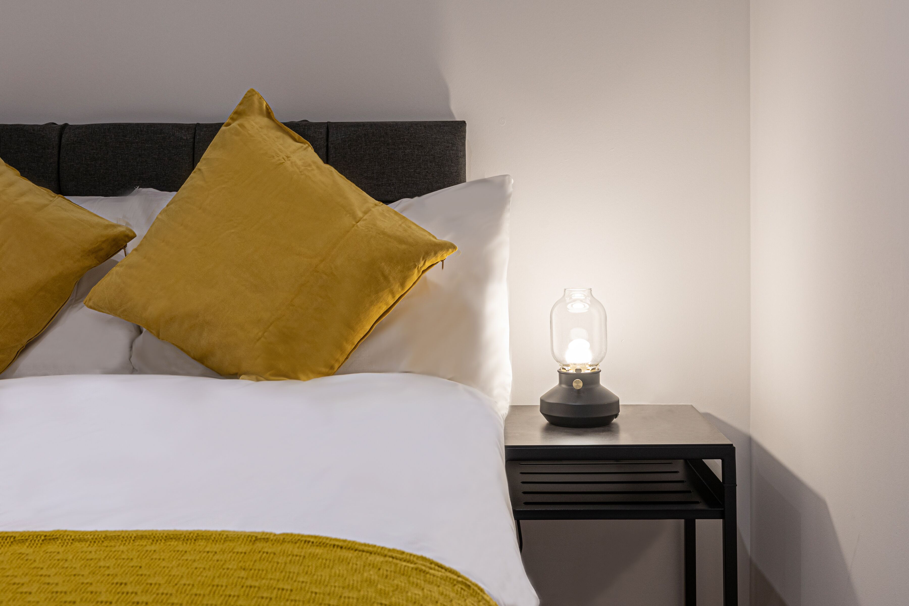 Un lit bien fait, agrémenté d’oreillers blancs et moutarde, une table de chevet avec une lampe et une tête de lit moderne.